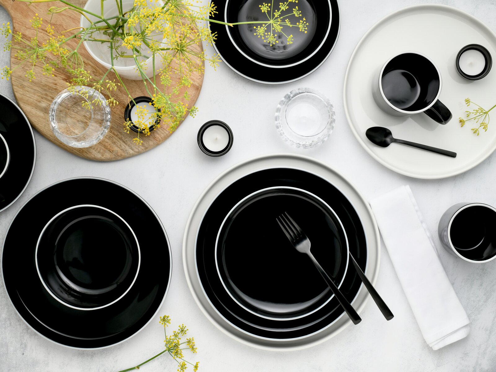 Draufsicht auf ein modern gedecktes Tisch-Arrangement mit schwarzem Geschirr mit weißem Rand, Tasse und Schalen, schwarzem Besteck, Teelichtern, Glas-Schälchen, Holzbrett und gelben Blüten auf hellem Hintergrund