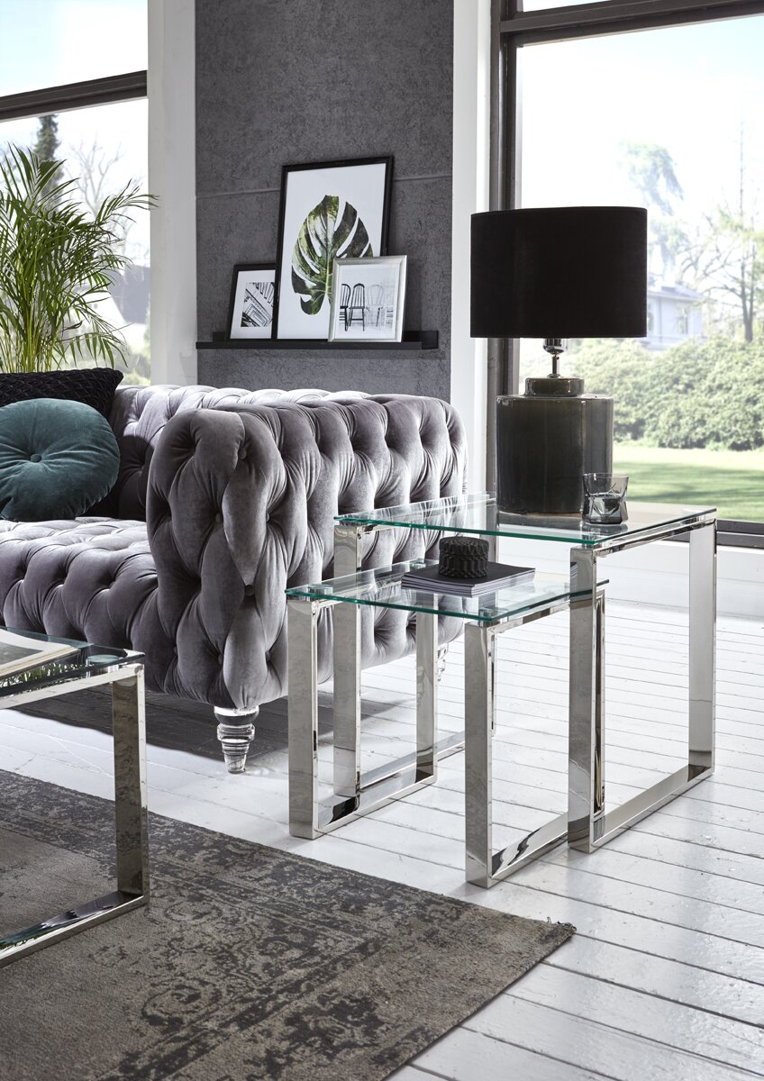 Zurbrüggen Beistelltisch Moderner Beistelltisch mit Glasplatten und verchromtem Metallgestell neben einem grauen Chesterfield-Sofa, Perspektive: leicht seitliche Ansicht auf Tischhöhe.
