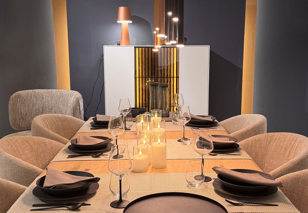 Gedeckter Tisch mit Weingläsern und vielen Kerzen in der Mitte, im Hintergrund steht eine Kommode, darauf eine Tischleuchte. 