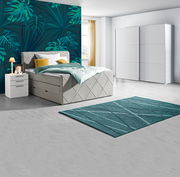 Modernes Schlafzimmer mit Boxspringbett in hellem Stoff, zwei weißen Nachttischen, einem großen weißen Schwebetürenschrank und einem türkisfarbenen Teppich, seitliche Perspektive.