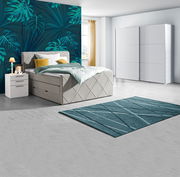 Modernes Schlafzimmer mit Boxspringbett in hellem Stoff, zwei weißen Nachttischen, einem großen weißen Schwebetürenschrank und einem türkisfarbenen Teppich, seitliche Perspektive.