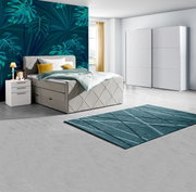 Modernes Schlafzimmer mit Boxspringbett in hellem Stoff, zwei weißen Nachttischen, einem großen weißen Schwebetürenschrank und einem türkisfarbenen Teppich, seitliche Perspektive.