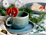 Hellgrüne Keramiktasse mit Kaffee auf blauem Unterteller, dekoriert mit herzförmigem roten Keks am Tassenrand; Perspektive: Nahaufnahme aus leicht erhöhter seitlicher Ansicht.