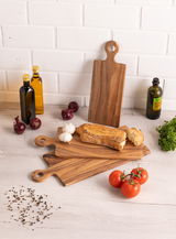Holzgriffbrett in einer Küche, umgeben von Olivenölflaschen, roten Zwiebeln, Knoblauch, Brot, Tomaten und Petersilie. Perspektive von oben.