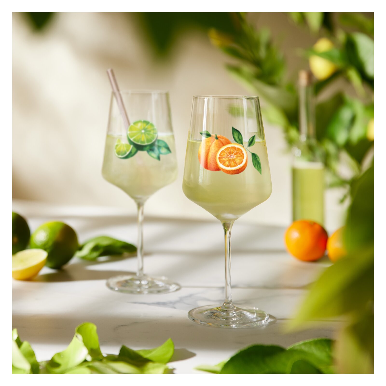 Zwei elegante Cocktailgläser mit frischem Zitrusdrink und Fruchtmotiven (Orange und Limette) auf hellem Tisch, Perspektive: frontale Nahaufnahme auf Augenhöhe mit leichtem Hintergrund-Bokeh.