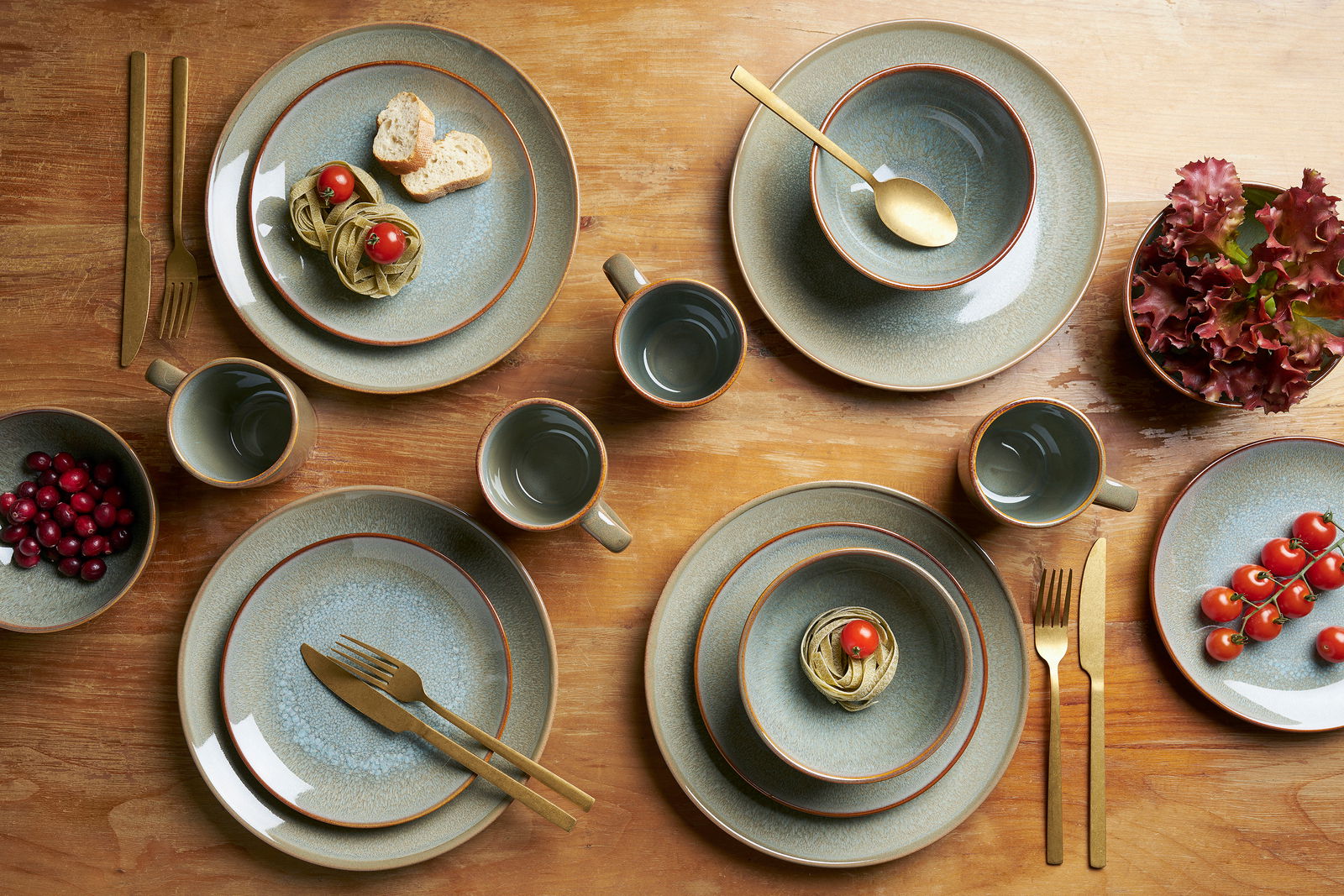 Vogelperspektive auf ein 16-teiliges Kombiservice-Set mit Tellern, Schalen und Tassen in erdigen Tönen, arrangiert auf einem Holztisch mit goldfarbenem Besteck und dekorativen Lebensmitteln.