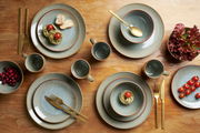 Vogelperspektive auf ein 16-teiliges Kombiservice-Set mit Tellern, Schalen und Tassen in erdigen Tönen, arrangiert auf einem Holztisch mit goldfarbenem Besteck und dekorativen Lebensmitteln.