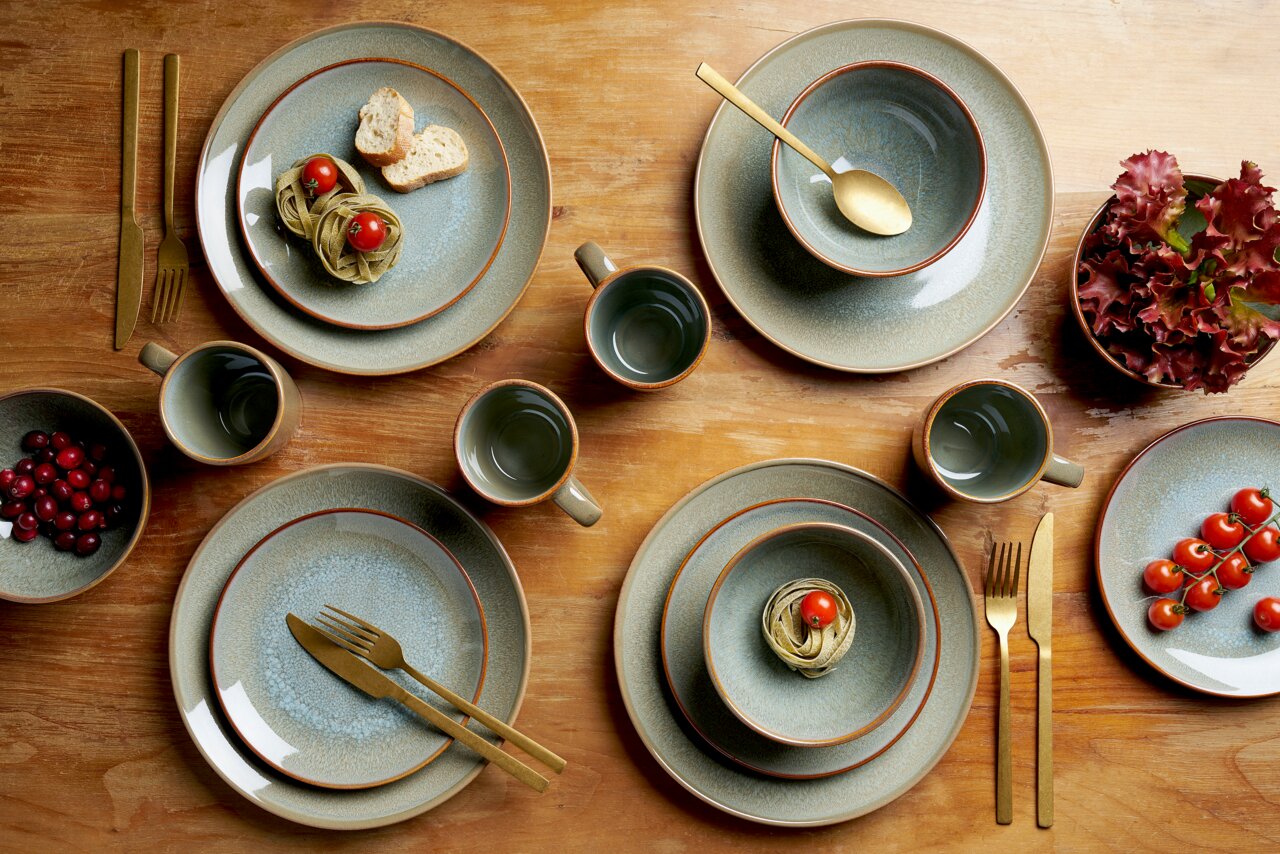 Vogelperspektive auf ein 16-teiliges Kombiservice-Set mit Tellern, Schalen und Tassen in erdigen Tönen, arrangiert auf einem Holztisch mit goldfarbenem Besteck und dekorativen Lebensmitteln.