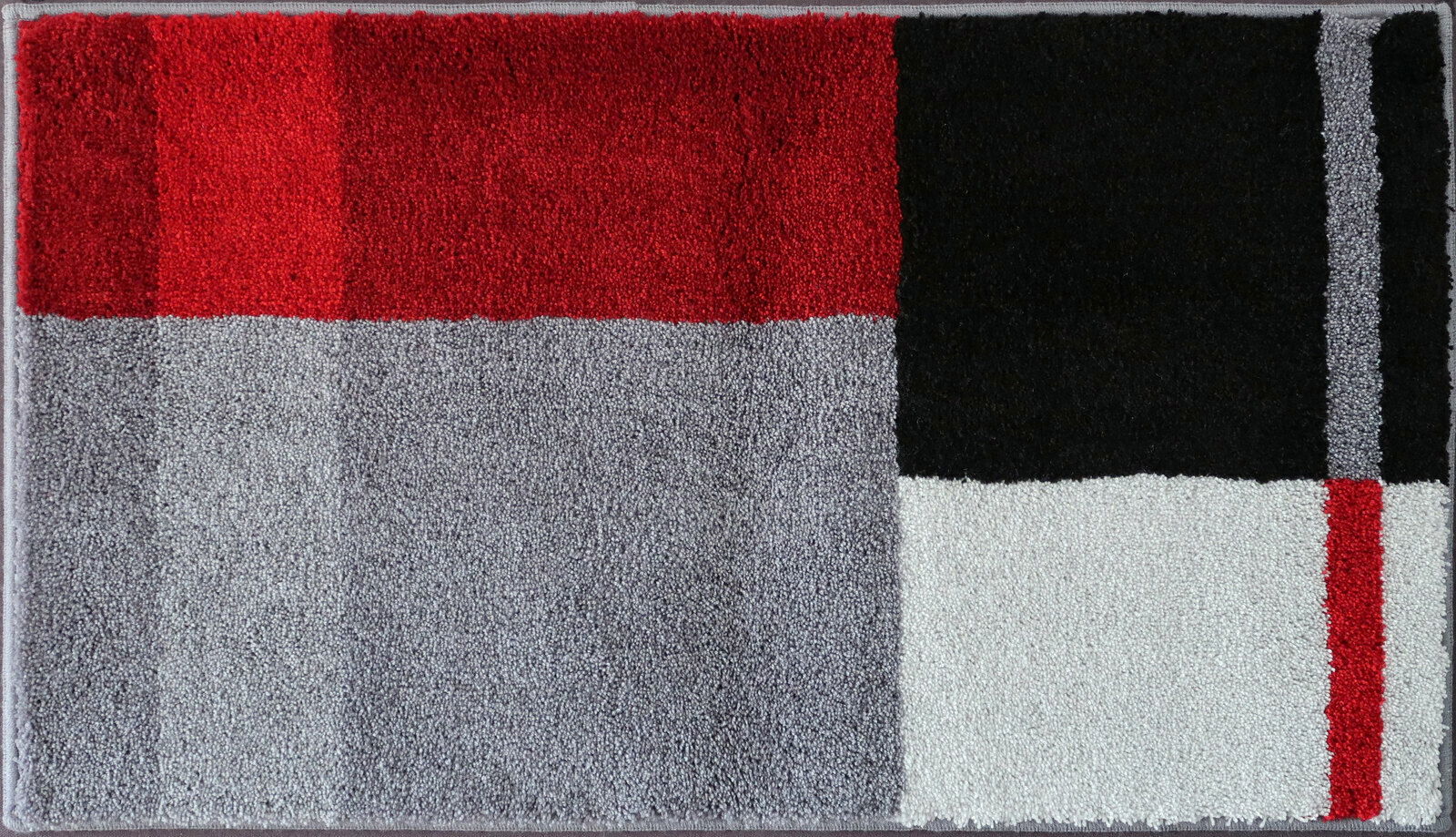 Badteppich in rechteckiger Form mit geometrischem Muster in Rot, Schwarz, Grau und Weiß, aus der Vogelperspektive fotografiert.