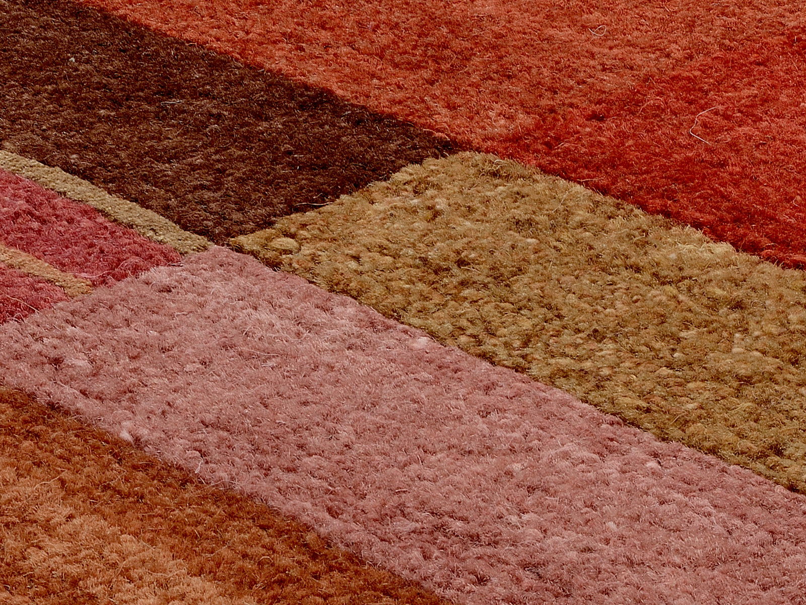 Nahaufnahme eines Wollteppichs mit verschiedenen rechteckigen Farbflächen in Rot-, Braun- und Beigetönen, aus einer schrägen Perspektive aufgenommen.