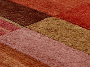 Nahaufnahme eines Wollteppichs mit verschiedenen rechteckigen Farbflächen in Rot-, Braun- und Beigetönen, aus einer schrägen Perspektive aufgenommen.