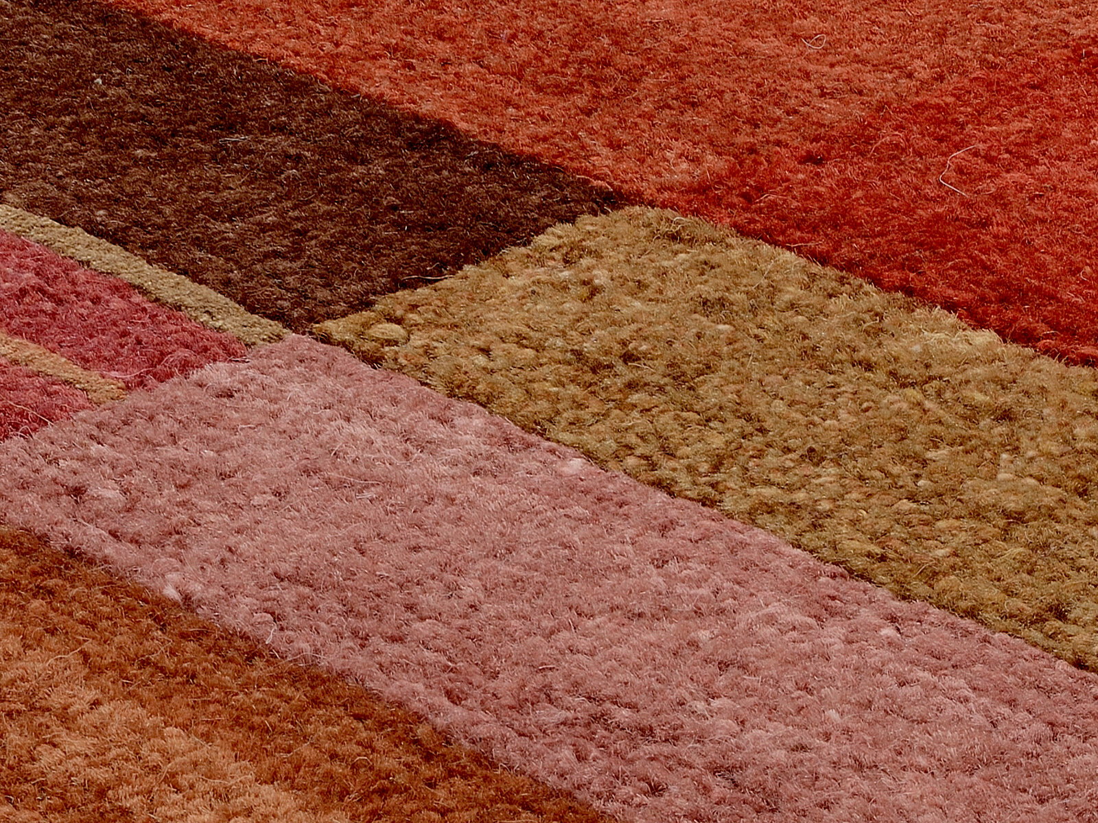 Nahaufnahme eines Wollteppichs mit verschiedenen rechteckigen Farbflächen in Rot-, Braun- und Beigetönen, aus einer schrägen Perspektive aufgenommen.