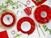 Draufsicht auf ein rot-weiß gepunktetes Geschirrset mit Tellern, Schalen und Tasse auf hellen Tischunterlagen, dekoriert mit goldfarbenem Besteck, Glas, Teelichtern, roten Servietten und blühenden Zweigen.