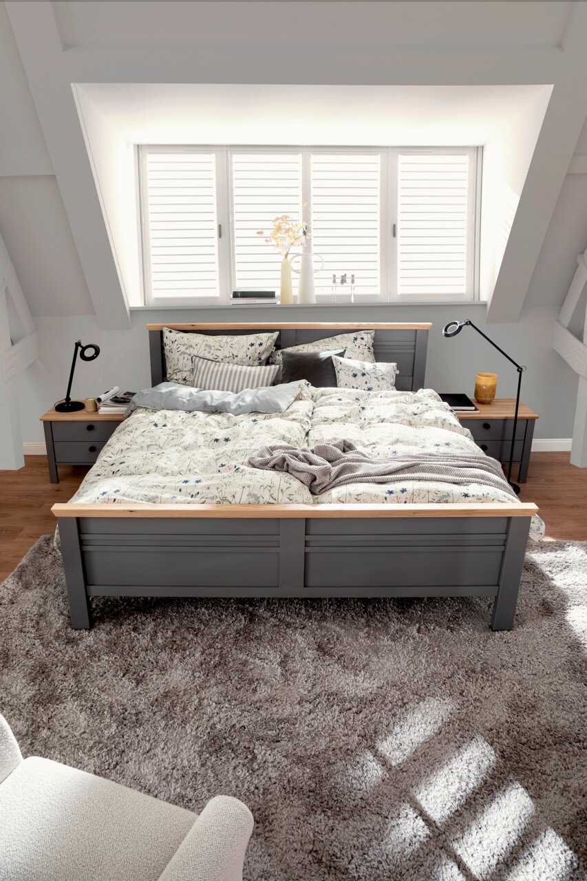 Een gezellige slaapkamer met een grijs houten bed, opgemaakt met bloemetjesbeddengoed in zachte kleuren. Het bed staat op een zacht tapijt, omringd door nachtkastjes met moderne lampen. Het perspectief toont het bed van voren, met op de achtergrond een ra