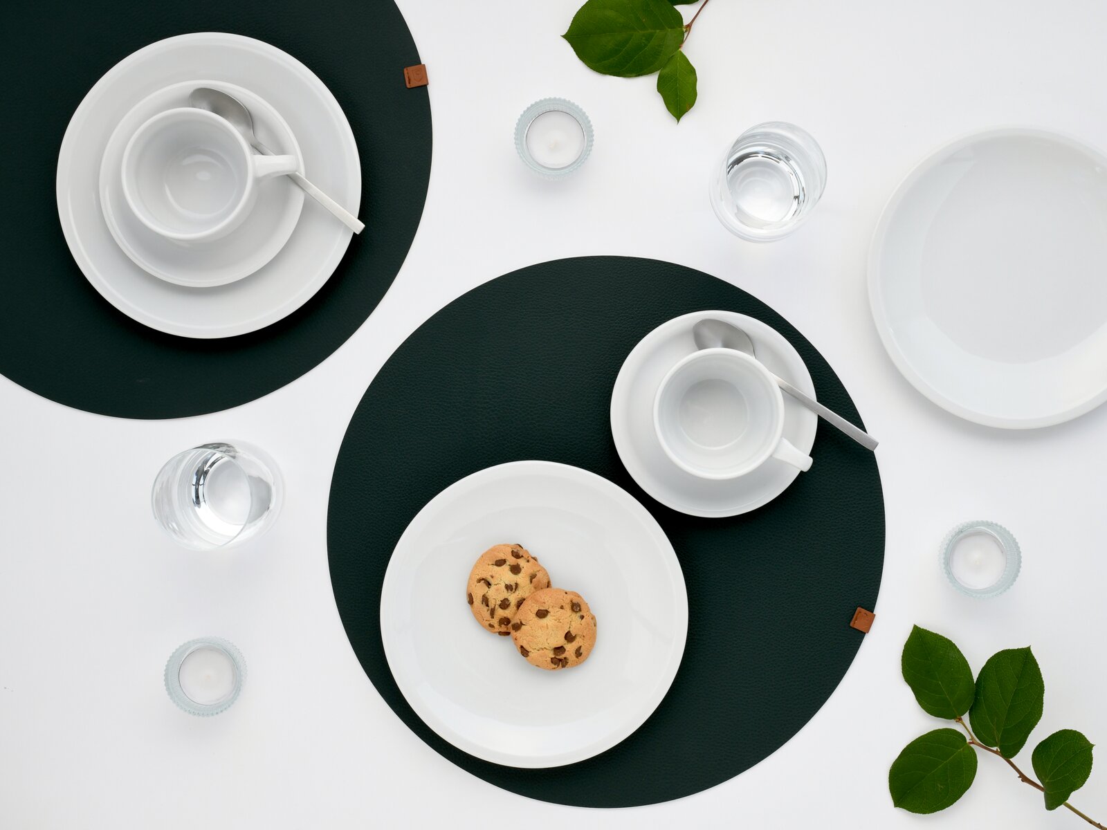 Draufsicht auf ein minimalistisches Tischgedeck mit weißen Tassen und Tellern auf dunkelgrünen Platzsets, Wassergläsern, Teelichtern und zwei Schokokeksen auf einem Teller