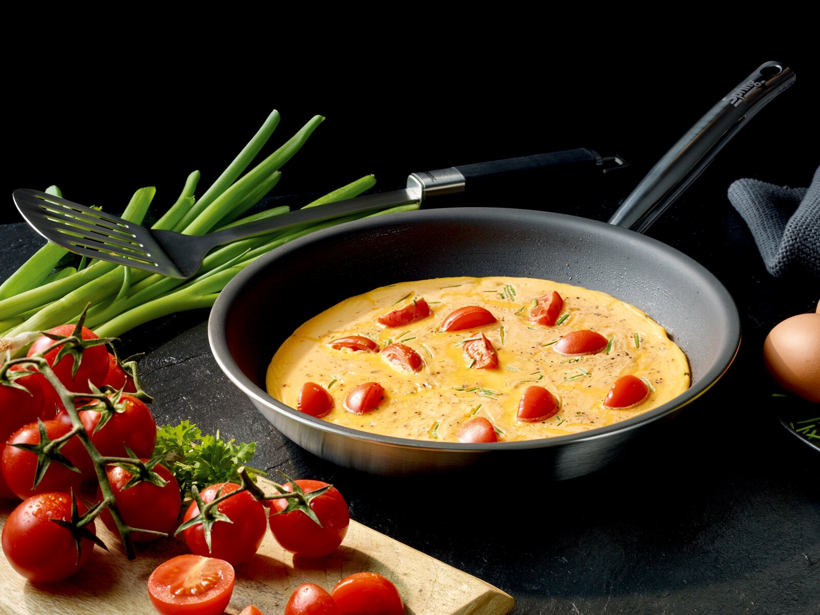 Spring Koekenpan XL 24 cm BRIGADE ULTIMATE Schuine bovenaanzichtfoto van een zwarte koekenpan met omelet/frittata met cherrytomaten en kruiden, omringd door lente-ui, tomaten op de tak, spatel en ei op donkere achtergrond