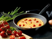 Schuine bovenaanzichtfoto van een zwarte koekenpan met omelet/frittata met cherrytomaten en kruiden, omringd door lente-ui, tomaten op de tak, spatel en ei op donkere achtergrond