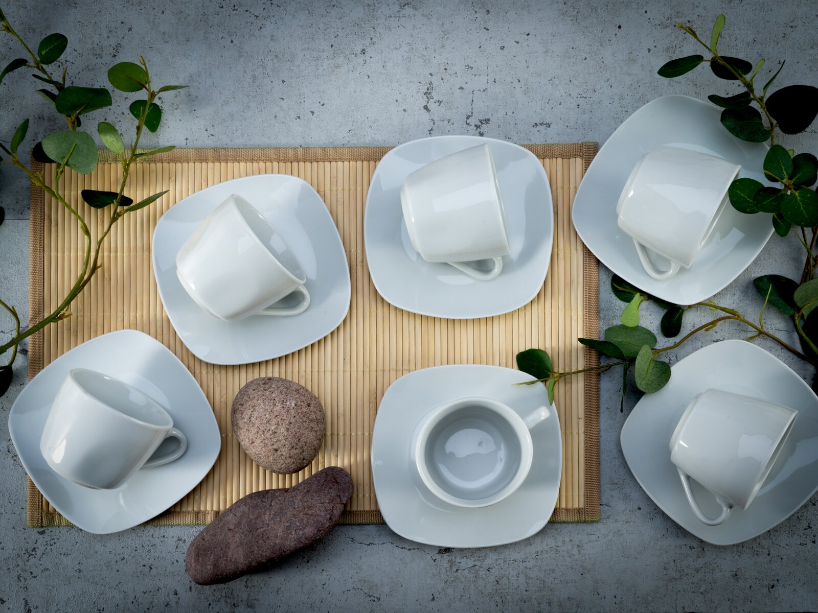 Fünf weiße Porzellan-Kaffeetassen mit passenden Untertassen im modernen, quadratisch abgerundeten Design auf Bambusmatte, dekoriert mit grünen Zweigen und Steinen, fotografiert aus der Vogelperspektive (Top-Down).