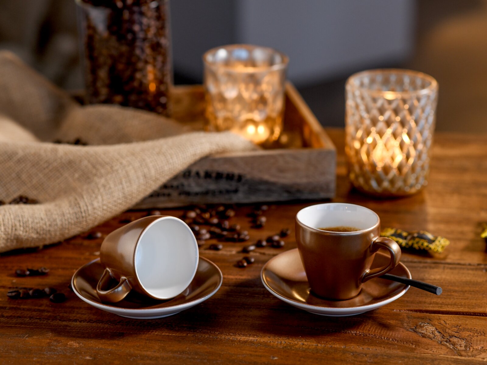 Creatable Espressoset 12-tlg GOLDEN DAYS Zwei braune Espressotassen mit Untertassen auf rustikalem Holztisch, eine Tasse liegend, im Hintergrund Kerzenlicht und Kaffeebohnen; Perspektive: leicht von vorn auf Tischhöhe.