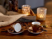 Zwei braune Espressotassen mit Untertassen auf rustikalem Holztisch, eine Tasse liegend, im Hintergrund Kerzenlicht und Kaffeebohnen; Perspektive: leicht von vorn auf Tischhöhe.