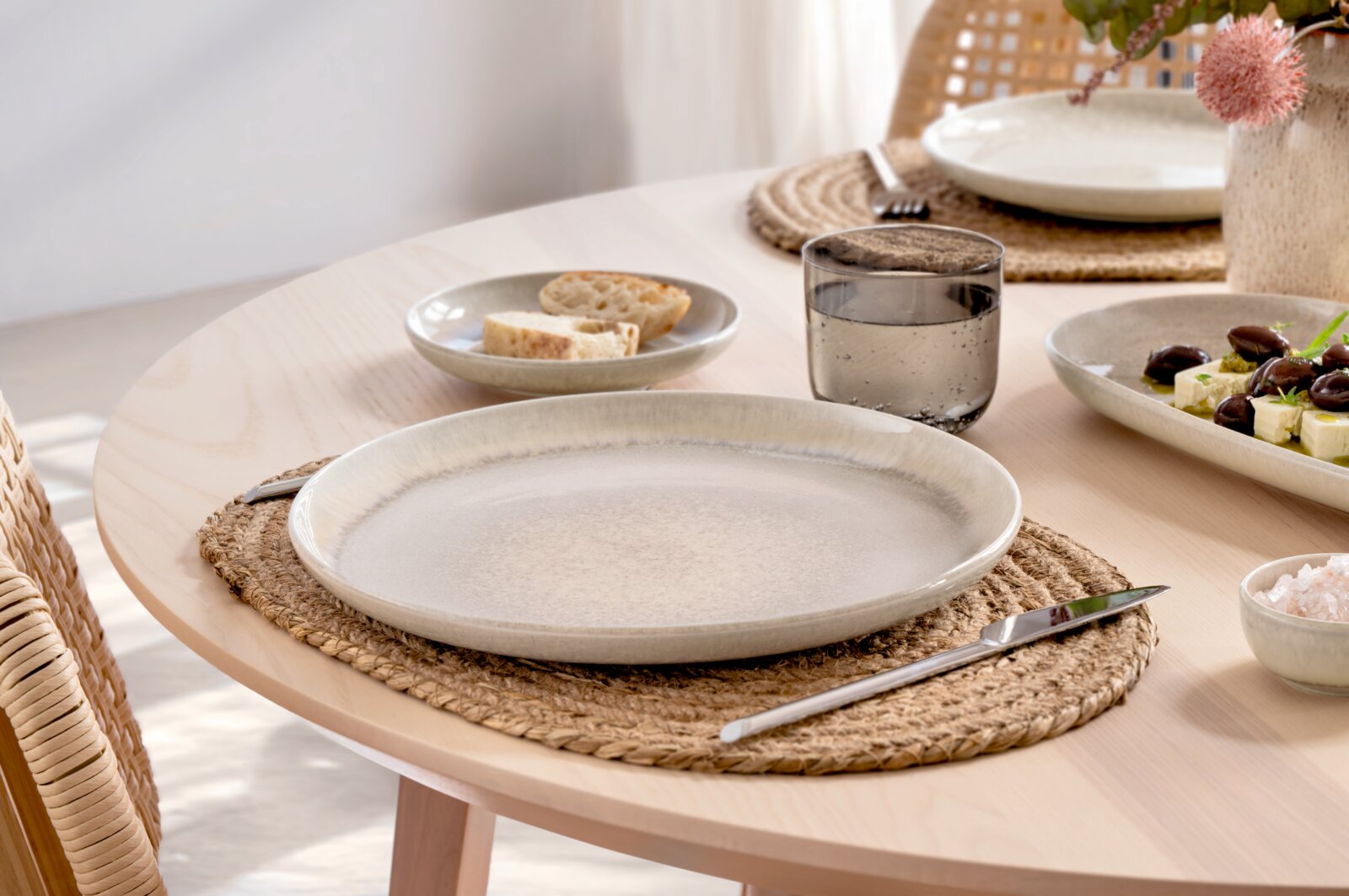 Beige keramisch dinerbord met subtiele randglazuur op gevlochten jute placemat op lichte houten eettafel, gefotografeerd in schuin vooraanzicht.