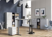 Moderne Büroumgebung mit höhenverstellbarem Schreibtisch, Rollcontainer und Aktenschränken in einem hellen Raum mit großen Fenstern, seitliche Perspektive.