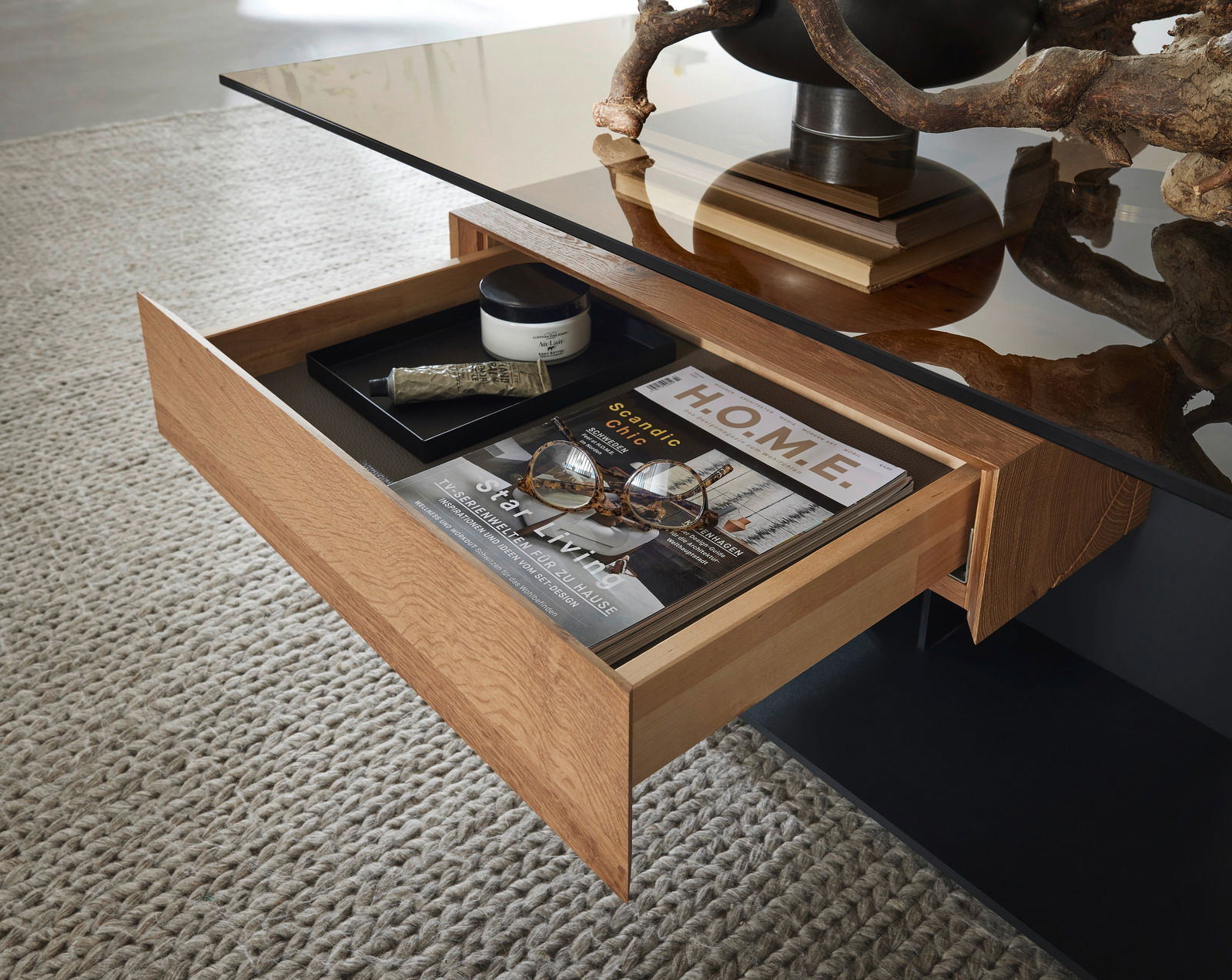 Moderner Couchtisch mit Glasplatte und offener Schublade, gefüllt mit Zeitschriften und Dekoration, aus der Perspektive von oben.