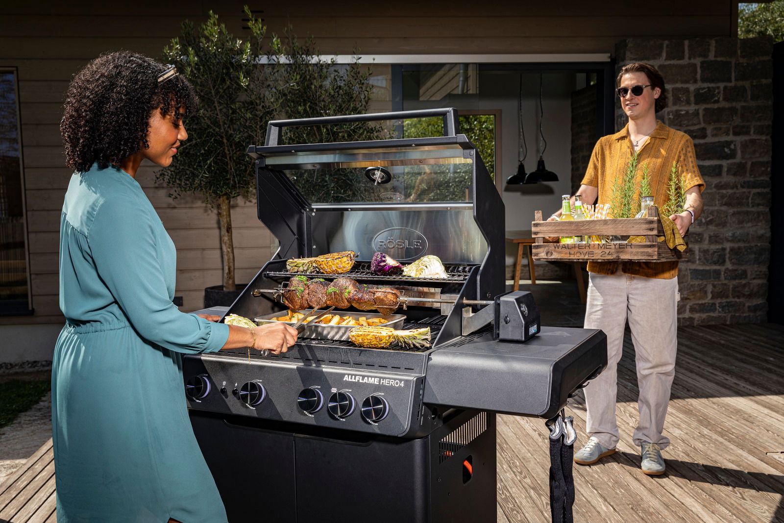 Zwei Personen beim Grillen mit dem Gasgrill Allflame Hero 4 auf einer Terrasse, eine Person grillt Gemüse und Fleisch, die andere bringt Getränke. Perspektive von der Seite des Grills.