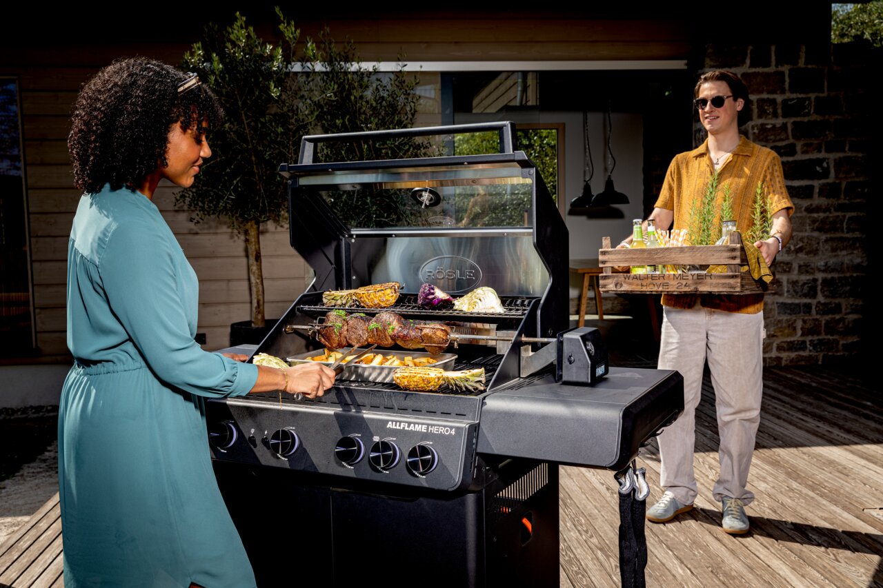 Twee mensen zijn aan het barbecueën met de Allflame Hero 4-gasbarbecue op een terras; de ene persoon grilt groenten en vlees, de andere brengt drankjes. Gezien vanaf de zijkant van de barbecue.