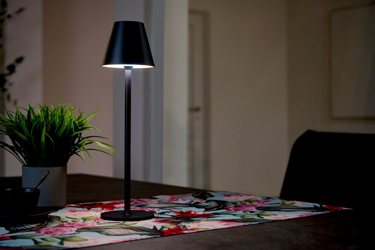 BOXXX Akku-Tischleuchte STEVE Schwarze Akku-Tischleuchte auf einem Tisch mit bunter Tischdecke, seitliche Perspektive