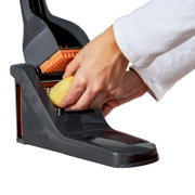 Close-up zijaanzicht van een hand die een geschilde aardappel door een grijs-oranje keukenmandoline met rooster snijdt op een witte achtergrond