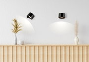 Zwei schwarze Decken-Aufbaustrahler in einem minimalistischen Raum, einer schwenkbar, mit warmem Lichtkegel an der weißen Wand; Perspektive: frontale Ansicht mit leichtem Blick nach oben.