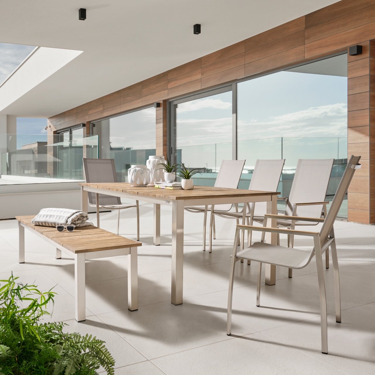 Moderne Outdoor-Essgruppe auf einer Terrasse: rechteckiger Tisch mit Holzplatte und Metallgestell, dazu helle Stühle und passende Bank, seitliche Frontansicht in leichter Perspektive