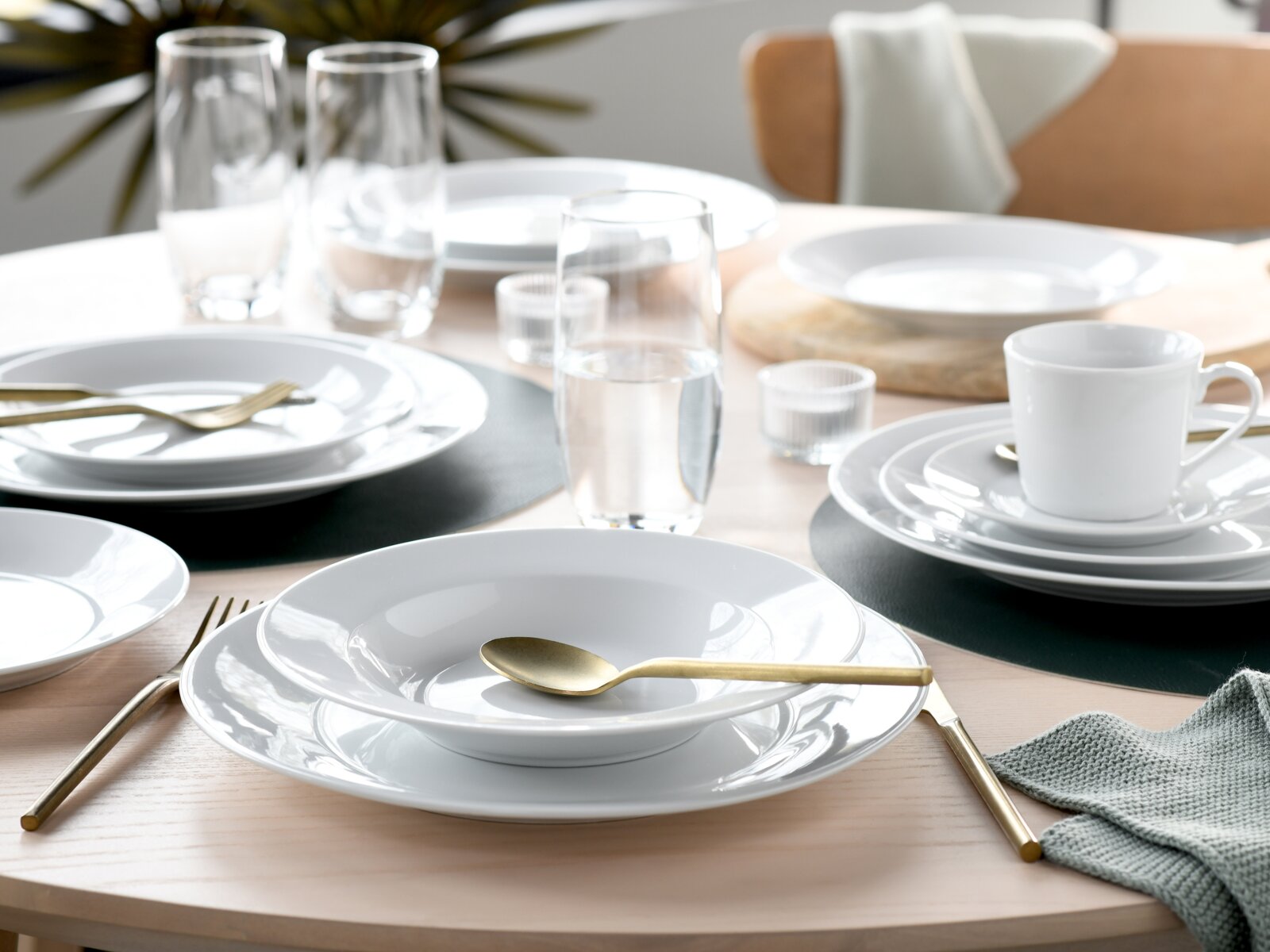 Modern gedeckter Esstisch mit weißem Porzellan-Geschirr (Teller und Schalen), goldfarbenem Besteck, klaren Wassergläsern und weißer Tasse auf dunklen Platzsets, fotografiert aus leicht erhöhter schräger Perspektive.