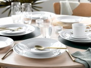 Modern gedeckter Esstisch mit weißem Porzellan-Geschirr (Teller und Schalen), goldfarbenem Besteck, klaren Wassergläsern und weißer Tasse auf dunklen Platzsets, fotografiert aus leicht erhöhter schräger Perspektive.