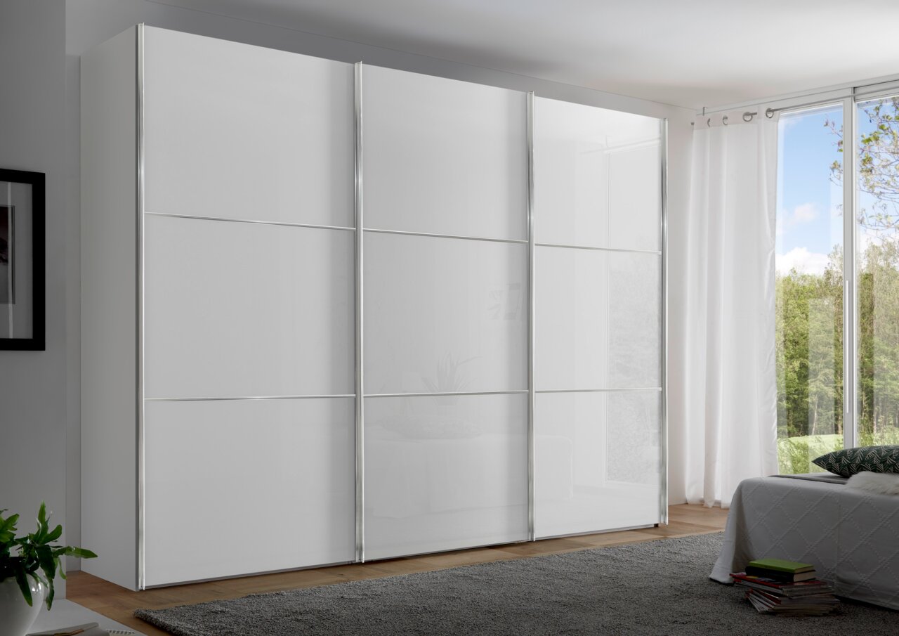 Moderner weißer Schwebetürenschrank mit drei Türen, seitliche Perspektive, in einem hellen Raum mit großen Fenstern.