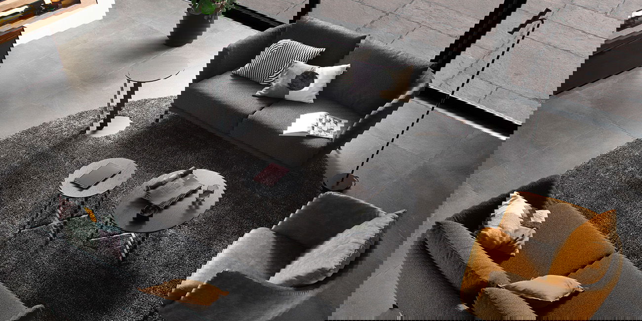 Ein stilvolles Wohnzimmer mit zwei grauen Sofas, einem gelben Sessel, und runden Tischen auf einem Plüschteppich.