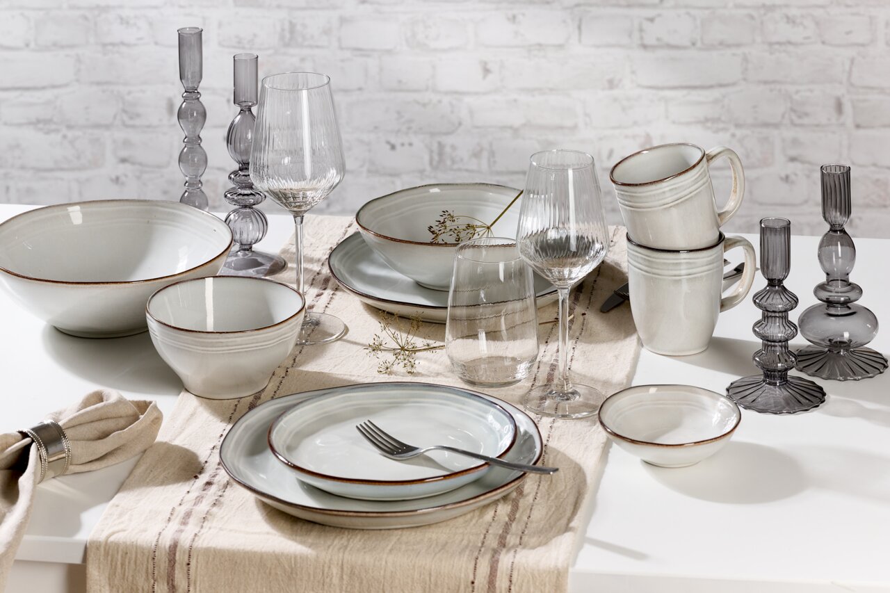 Frühstücksset 12-tlg. aus Keramik in neutralen Tönen, bestehend aus Tellern, Schalen und Tassen, auf einem Tisch mit Tischläufer und Weingläsern, seitliche Perspektive.
