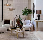 Frontalansicht eines stilvoll eingerichteten Wohnbereichs mit cremefarbenen Lounge-Sesseln, eleganten Messing-Couchtischen, skulpturalen Pendelleuchten und üppigen Grünpflanzen.