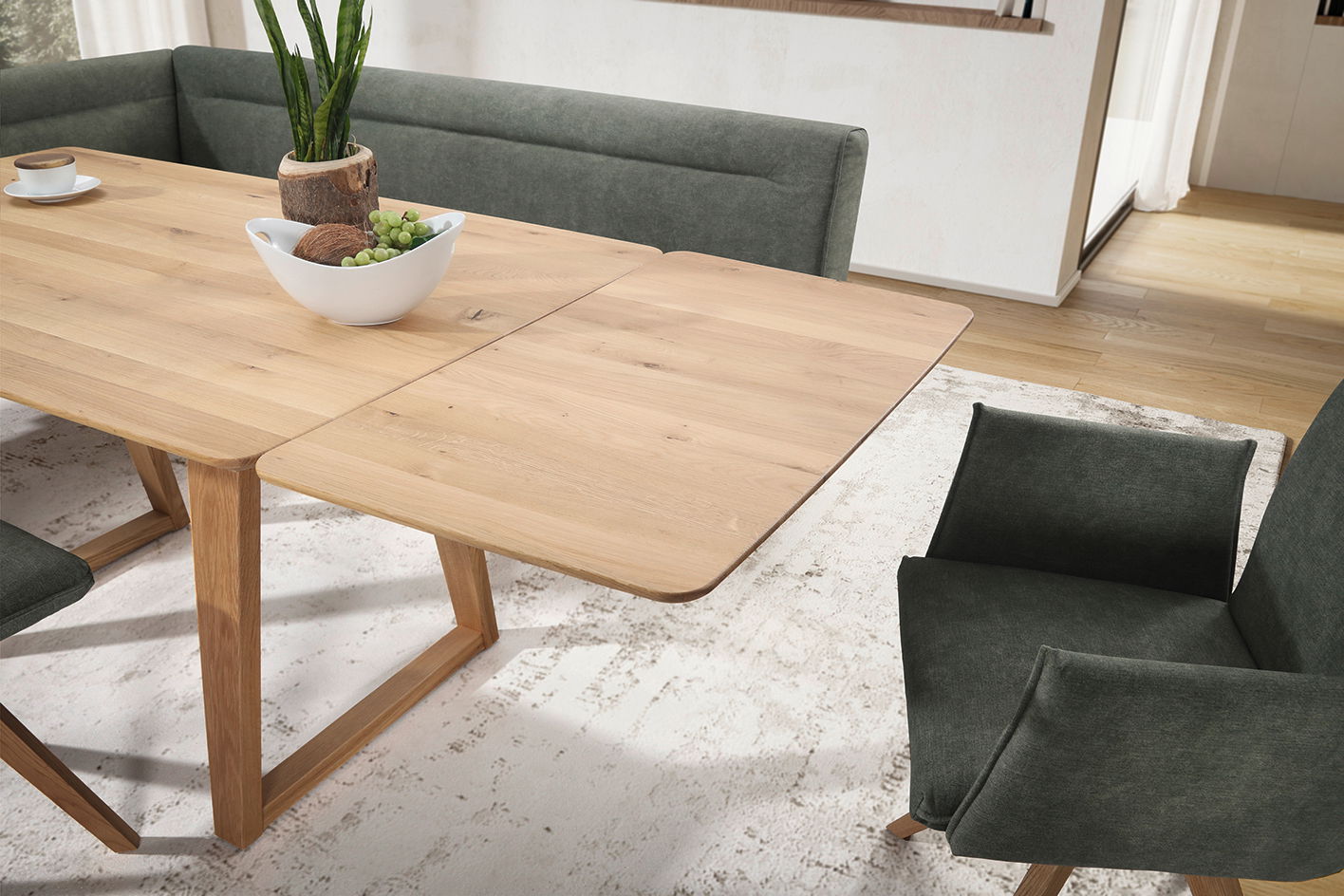 Esszimmer mit einem ausziehbaren Holztisch und grünen Polsterstühlen, Perspektive von schräg oben.