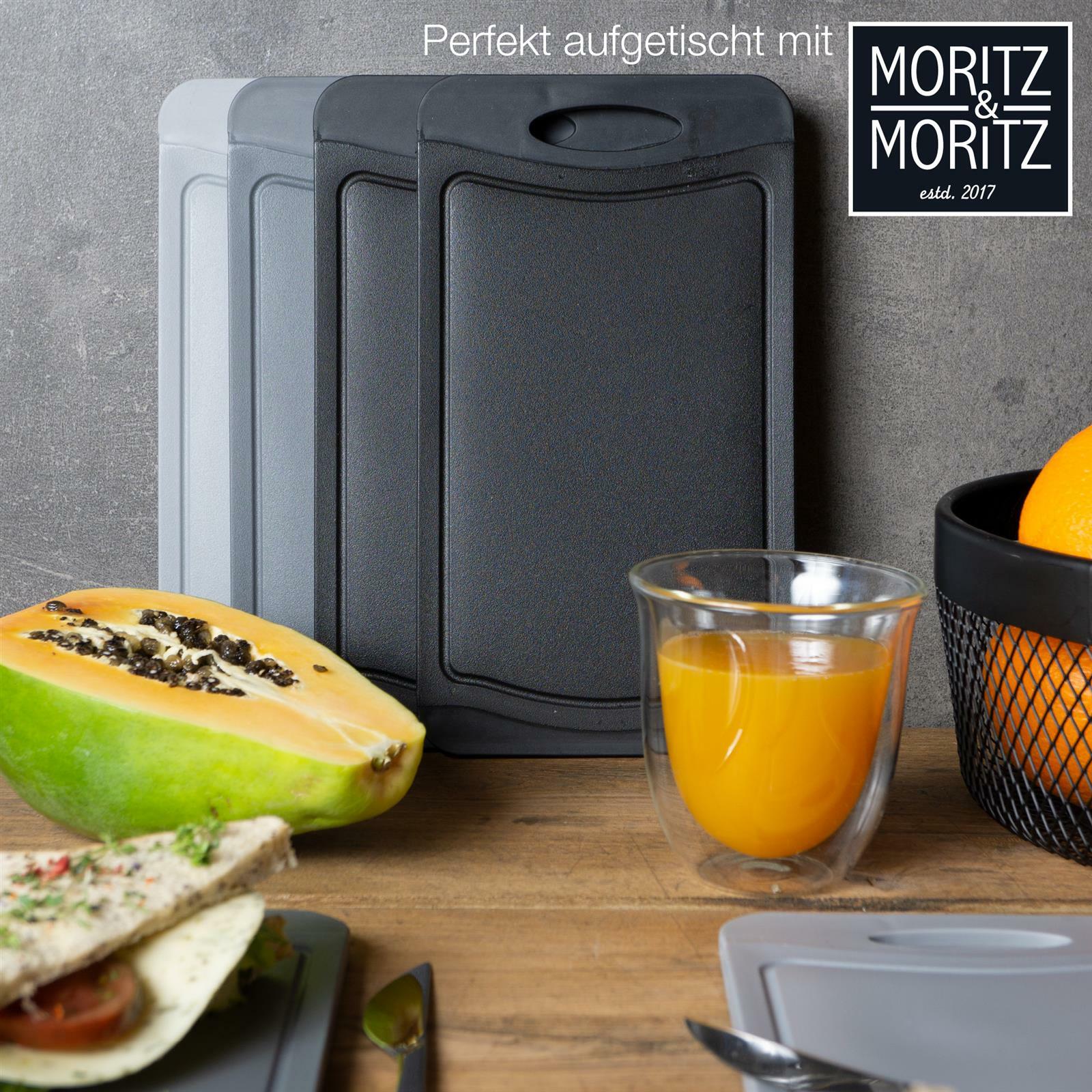 Drei Schneidebretter in verschiedenen Grautönen stehen aufrecht auf einem Holztisch. Im Vordergrund sind ein halbierte Papaya, ein Glas Orangensaft und ein Teller mit einem belegten Wrap zu sehen. Perspektive: seitlich.