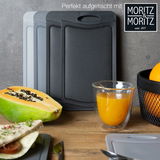 Drei Schneidebretter in verschiedenen Grautönen stehen aufrecht auf einem Holztisch. Im Vordergrund sind ein halbierte Papaya, ein Glas Orangensaft und ein Teller mit einem belegten Wrap zu sehen. Perspektive: seitlich.