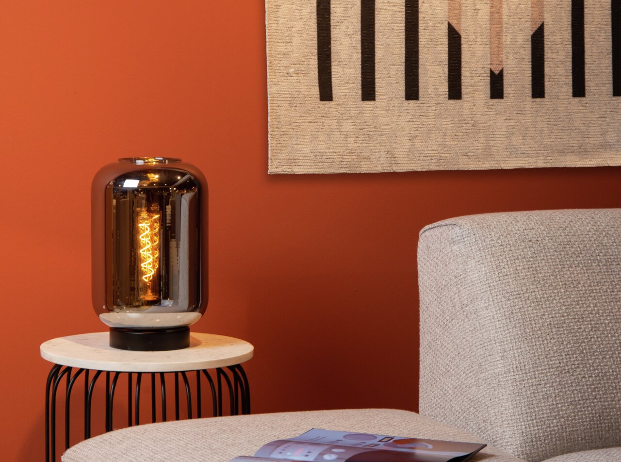 Tafellamp JOANET met rookgrijs glas op een rond bijzettafeltje, zijaanzicht, tegen een oranje muur.