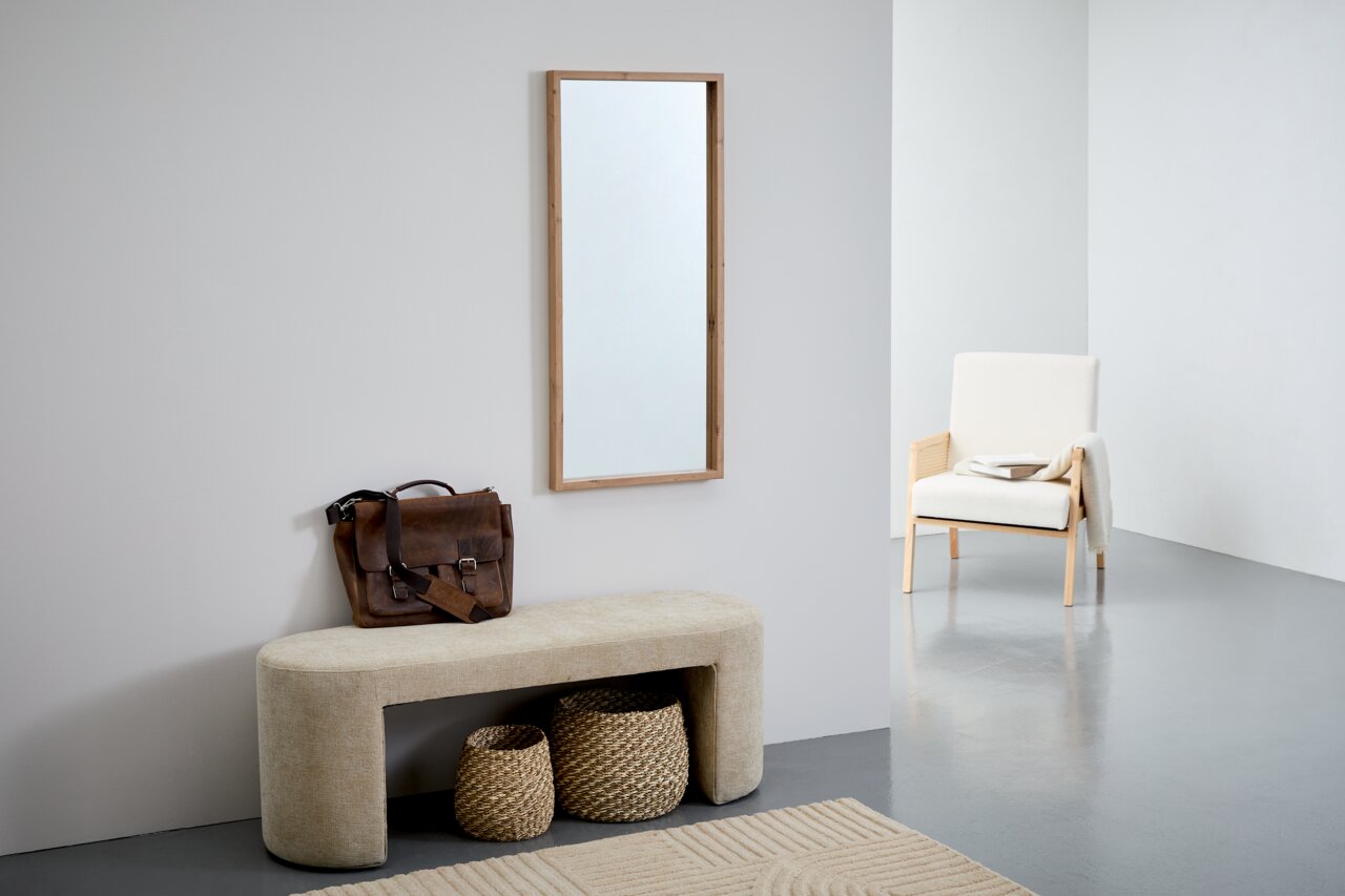 Rechteckiger Spiegel mit Holzrahmen, an einer hellen Wand hängend, im modernen Wohnambiente mit Bank, Korb und Sessel. Perspektive von vorne.
