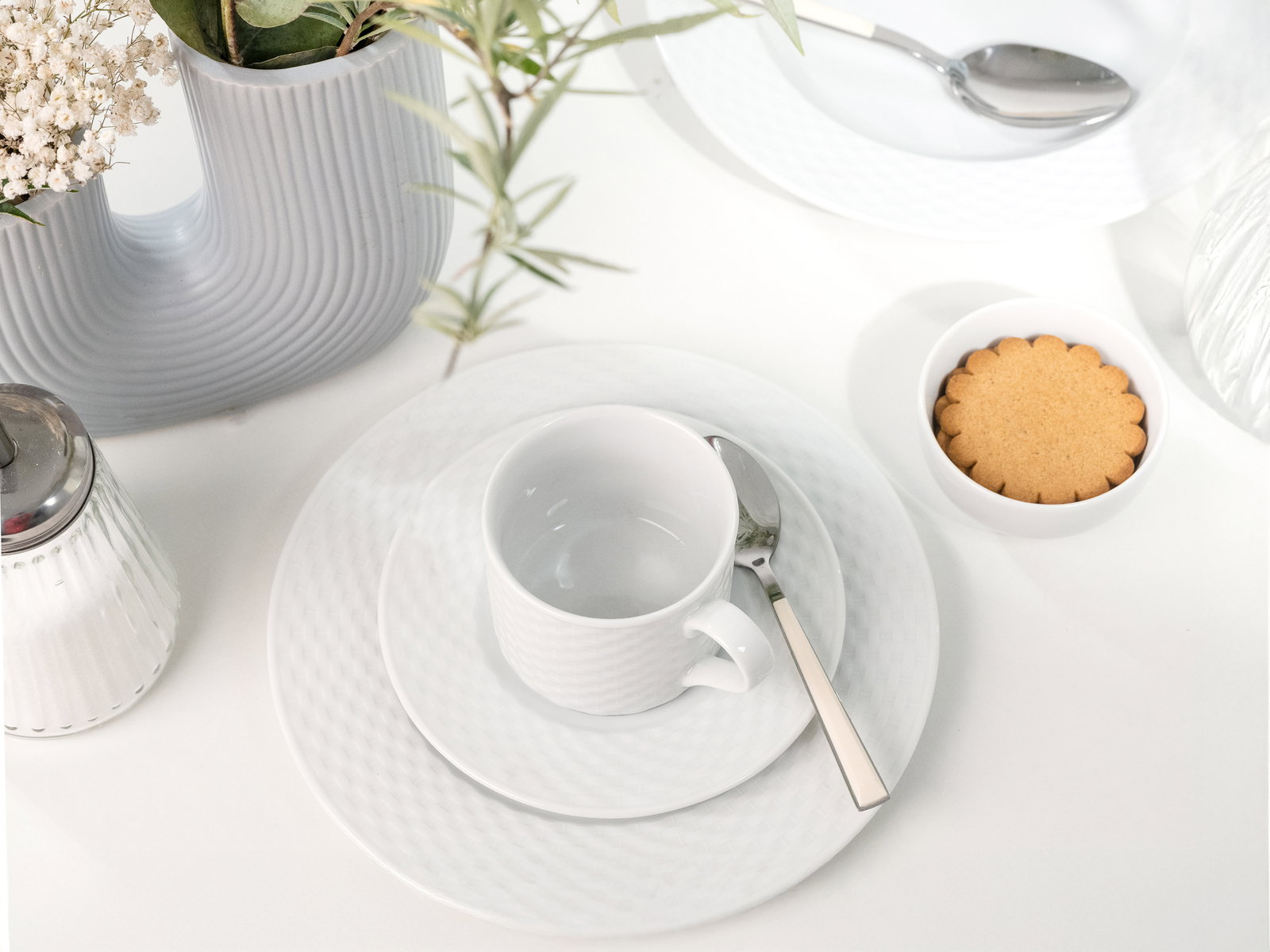 Kaffeeservice mit Tasse, Untertasse und Teller in eleganter weißer Keramik, von oben fotografiert, neben einem Keks und einer dekorativen Vase.
