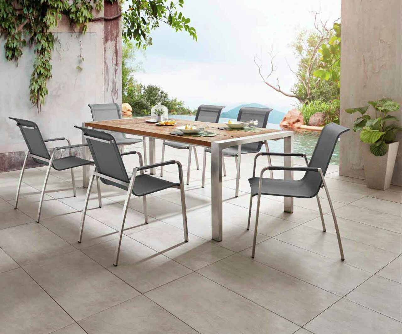 Moderner Gartentisch mit Holzplatte und Metallbeinen, umgeben von sechs stilvollen Stühlen, auf einer Terrasse mit Blick auf einen Pool und üppige Vegetation, seitliche Perspektive.