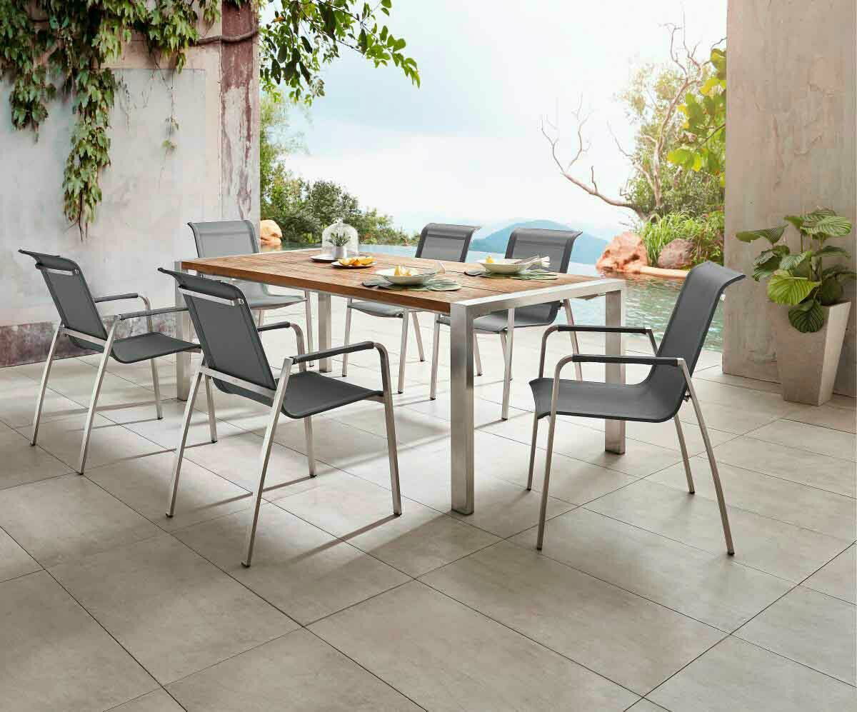 Moderner Gartentisch mit Holzplatte und Metallbeinen, umgeben von sechs stilvollen Stühlen, auf einer Terrasse mit Blick auf einen Pool und üppige Vegetation, seitliche Perspektive.