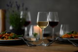 Ein stilvolles Wasser- und Rotweinglas Classico auf einem gedeckten Tisch, frontal fotografiert, umgeben von Tellern mit Pasta und weiteren Weingläsern im Hintergrund.