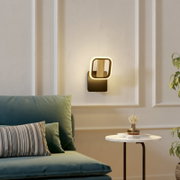 Moderne LED-Wandleuchte mit warmweiß leuchtendem, abgerundet-quadratischem Rahmen, schwarzer Wandhalterung und Holzelement, frontal in leicht seitlicher Perspektive im Wohnzimmer an heller Wand montiert