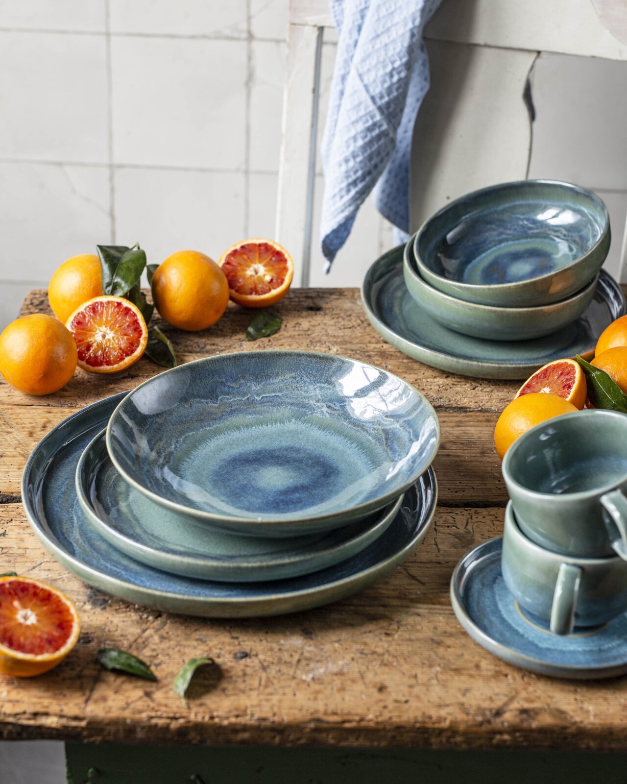 Blaugrünes, glasiertes Keramik-Geschirrset mit Tellern, Schalen und Tassen auf rustikalem Holztisch, dekoriert mit Blutorangen Perspektive: leicht erhöhte Frontansicht.
